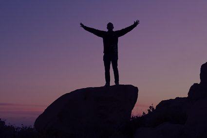 Silhouette of person with out stretched arms standing on a rock at dusk