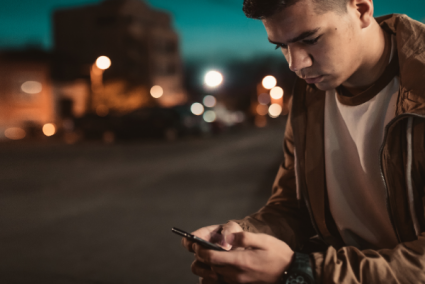 A teenager looking at their smart phone on a street at night time.